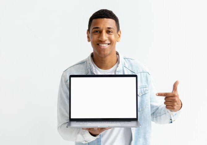 Portrait of young African American man with laptop on white background. Male points her finger at a blank laptop screen, looking at the camera and smiling